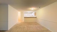 This single-floor space features an open-concept layout that connects the living area to the kitchen, creating a cohesive and functional flow. The living area is finished with wall-to-wall beige carpeting, which adds a sense of warmth underfoot, while the neutral cream-toned walls provide a clean, versatile backdrop throughout. Overhead flush-mount lighting illuminates the space evenly.

The kitchen is separated from the living area by a half-wall peninsula, which serves as a natural divider while maintaining visual openness. White cabinetry lines the kitchen walls, offering ample storage, and the countertops complement the light, airy aesthetic of the space. A doorway visible to the side suggests additional rooms or hallways branching off from the main living area, contributing to the overall layout's practicality.

The textured ceiling adds subtle dimension, and the consistent neutral palette throughout creates a cohesive, move-in-ready environment. The open connection between the kitchen and living area makes this space well-suited for everyday living and casual entertaining.