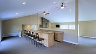 This single-floor space is thoughtfully designed for entertaining and social gatherings, offering an open and inviting atmosphere throughout. The centerpiece of the room is a generously sized bar with light wood cabinetry and granite countertops, lined with a row of elegant wrought-iron bar stools that provide ample seating for guests. Below the bar, built-in cabinetry and an under-counter appliance offer practical storage and convenience for hosting.

The room's vaulted ceilings lend a sense of grandeur and openness, further enhanced by dual ceiling fans with integrated lighting that ensure comfortable air circulation. Recessed lighting throughout the ceiling adds a warm, even glow to the space. A striking stone fireplace serves as a dramatic focal point at the far end of the room, adding a rustic yet refined character to the overall design.

Natural light fills the space through a series of windows along the exterior wall, offering pleasant views of the surrounding wooded landscape. A wall-mounted flat-screen television provides additional entertainment options. Neutral-toned carpeting and warm beige walls create a welcoming backdrop, while a pass-through opening connects to an adjacent area, ensuring seamless flow and versatility throughout the layout.