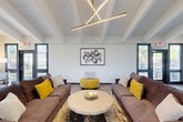 Located at 2100 Thicket Pl SE in Huntsville, Alabama, this thoughtfully designed community space delivers a striking blend of modern aesthetics and welcoming comfort. The single-floor layout makes efficient use of its footprint, anchored by a dramatic vaulted ceiling with exposed white-painted beams that lend an airy, open feel throughout.

The primary lounge area is a standout, featuring two large brown sofas accented with bold mustard yellow and neutral throw pillows, arranged around a round marble-top coffee table. A sculptural linear pendant light fixture crowns the space, while a framed abstract artwork adorns the central wall. Black-framed glass doors and windows on both sides of the lounge flood the room with natural light and offer views of the surrounding greenery.

Adjacent to the lounge, a more casual seating zone features curved accent chairs and a dining table with wooden chairs, all positioned beneath a large angular window wall that frames outdoor tree views beautifully. A ceiling fan adds practical comfort to this bright gathering area.

The kitchenette is well-appointed with white shaker cabinetry, granite countertops, a central island with bar seating, and black appliances including a refrigerator, microwave, and blender. A staircase is visible in the background, and interior clerestory windows add architectural interest while connecting the upper and lower visual zones of the space.