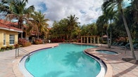 This inviting outdoor space centers around a generously sized, freeform resort-style swimming pool with crystal-clear turquoise water, accented by classic blue tile detailing along its curved edges. An attached spa seamlessly adjoins the main pool, offering a relaxing retreat within the same elegant setting. The pool deck is finished in warm terracotta-toned pavers, providing a comfortable and visually appealing surround. The landscape design is a standout feature, with tall, mature palm trees and a rich variety of tropical foliage framing the entire area, creating a private, lush atmosphere reminiscent of a Caribbean getaway. Vibrant red and green plantings add color and depth to the greenery throughout. A classic pergola with decorative columns anchors one side of the space, offering a shaded gathering area that blends seamlessly with the tropical surroundings. A wrought iron security fence encloses the area, ensuring both safety and a well-defined boundary. The overall ambiance is that of a well-maintained, resort-caliber outdoor amenity space — bright, open, and designed to maximize the enjoyment of a warm, sunny climate.