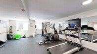Located at 9300 W Mall Dr in Everett, Washington, this fitness center offers a well-organized, single-room workout facility designed to accommodate a range of exercise needs. The space features durable rubber flooring throughout, providing a safe and practical surface for all types of training. Recessed and flush-mounted ceiling lights keep the room well-lit, while several windows along one wall allow natural light to filter in, creating a bright and inviting atmosphere. A large wall-mounted mirror spans one side of the room, visually expanding the space and allowing users to monitor their form during workouts. The cardio section is equipped with treadmills, an elliptical trainer, and a stationary bike, while a multi-station cable machine anchors the strength training area. A dumbbell rack, weight plates, and a stability ball round out the free weights section, catering to varied fitness routines. Posted exercise charts on the walls serve as handy workout references. A Coca-Cola vending machine near the entrance provides convenient access to beverages. The facility is accessed through a standard entry door and is maintained with a clean, neutral color palette of white and light gray, contributing to a focused and functional workout environment.