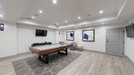 Located at 1533 Ellis Rd in Durham, NC, this single-floor entertainment and lounge space offers a well-appointed retreat designed for socializing and recreation. The room features light wood-look luxury vinyl flooring, crisp white walls, and a tray ceiling outfitted with recessed lighting that creates a bright, clean ambiance throughout.

At the heart of the space sits a solid wood pool table positioned atop a geometric-patterned area rug, serving as the room's central focal point. A shiplap accent wall with a wall-mounted flat-screen TV anchors one end of the room, while additional wall-mounted TVs provide entertainment from multiple vantage points. In-ceiling speakers further enhance the audio-visual experience.

A seating lounge area features a pair of plush tufted swivel chairs paired with chic gold-legged marble-top side tables, offering a comfortable spot to relax between games. A high-top bistro table with leather stools provides additional casual seating. The walls are adorned with bold, large-format abstract artwork in navy and white tones, lending a gallery-like sophistication to the space.

Wall sconces complement the recessed lighting, adding warmth to the modern aesthetic. The space is accessible via a glass-panel entry door and includes a secondary interior door, making it a versatile and fully self-contained entertainment venue.