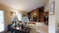 Located at 9696 Hayne Blvd in New Orleans, Louisiana, this single-story home offers a comfortable and inviting layout designed for easy living. The living room serves as the heart of the home, showcasing a striking floor-to-ceiling wood-paneled accent wall that frames a classic fireplace, creating a warm and cozy focal point. Vaulted ceilings with recessed lighting add a sense of openness and airiness to the space, while large windows allow natural light to filter in. Plush carpeting throughout the living area contributes to the relaxed, homey atmosphere. Built-in shelving alongside the fireplace provides practical display and storage space. The layout flows naturally toward a dining area, visible to the side, hinting at a well-connected floor plan suited for both everyday living and entertaining. The warm wood tones, neutral wall colors, and soft furnishings come together to create an atmosphere that feels lived-in and welcoming. This New Orleans home combines classic architectural details with functional design, making it a distinctive single-floor residence on the east side of the city.