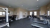 This fitness center offers a clean, functional environment purpose-built for exercise and physical wellness. The space features durable dark rubber flooring throughout, providing a safe and resilient surface suited to a wide variety of workouts. Recessed lighting in the smooth ceiling ensures the room is well-lit, complemented by natural light streaming in through windows along the far wall.

The room is thoughtfully arranged with a diverse selection of equipment. A tiered dumbbell rack lines one wall, stocked with a full range of free weights for strength training. A multi-station cable machine occupies a central position, offering versatility for resistance-based exercises. A SportsArt treadmill is prominently positioned for cardiovascular training, and additional equipment such as a weight bench and stability ball rounds out the offerings.

Motivational wall lettering adds an energizing touch to the neutral beige walls, fostering a focused workout atmosphere. A water fountain is conveniently located near the entry door, ensuring easy access to hydration. The overall layout is open and uncluttered, allowing ample space for movement and exercise. This fitness center is well-suited for a comprehensive training regimen in a comfortable, organized setting.