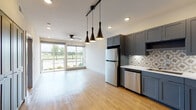 This contemporary single-floor residence offers a thoughtfully designed open-concept layout that seamlessly connects the kitchen and living area. The kitchen is a standout feature, showcasing gray shaker-style cabinetry, stainless steel appliances including a refrigerator, range, microwave, and dishwasher, and a decorative patterned tile backsplash. A trio of sleek pendant lights on a black track rail adds a modern industrial touch above the kitchen and dining area, complementing the recessed lighting throughout. Light wood-tone flooring flows continuously through the main living space, enhancing the bright and airy atmosphere. Large sliding glass doors and windows flood the living area with natural light and offer views of the surrounding greenery. A ceiling fan serves both the kitchen-adjacent living space and the bedroom areas. The bedroom features plush carpeting, a ceiling fan, and a window with outdoor views. The bathroom is well-appointed with gray vanity cabinetry, a white countertop, a tub-shower combination with tiled surround, and direct access to a spacious walk-in closet with built-in shelving. A wide, light-filled hallway with bold dark-paneled closet doors adds architectural character and practical storage to the unit's interior.