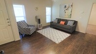 This single-floor space offers a comfortable and stylish living environment with a thoughtfully designed layout. The living area is a standout feature, showcasing rich, dark hardwood-style flooring that anchors the room with warmth and visual depth. Multiple windows allow generous natural light to fill the space, creating a bright and welcoming atmosphere throughout the day.

The living room is designed with a modern aesthetic, centered around a plush gray sofa accented with bold chevron and solid-colored throw pillows, complemented by a matching patterned accent chair. A soft, textured area rug in a trellis pattern grounds the seating arrangement and adds a layer of comfort underfoot. Decorative wall art adds a vibrant, artistic touch, lending personality and character to the room.

Additional details such as a sleek floor lamp and compact shelving unit contribute to the functional yet stylish nature of the space. The neutral wall tones provide a versatile backdrop that allows furnishings and décor to stand out. The overall ambiance is modern, cozy, and inviting, making it well-suited for both relaxing and entertaining. The single-floor layout ensures easy accessibility and a seamless flow throughout the entire living environment.
