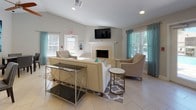 This single-floor common area is designed as a welcoming and functional gathering space, combining an open-concept layout with thoughtful amenities throughout. The main lounge area is anchored by a stone-surround fireplace with a white mantel, above which a wall-mounted flat-screen TV is positioned for easy viewing. Comfortable cream-toned upholstered seating, including a sofa and accent chairs with nail-head detailing, is arranged around a geometric-patterned area rug, creating an inviting atmosphere. Vaulted ceilings in this area add a sense of openness and volume.

The kitchenette is outfitted with warm wood cabinetry, a stainless steel refrigerator with an in-door ice and water dispenser, a microwave, and a dark granite-topped breakfast bar with stylish upholstered bar stools. Teal curtains frame the windows throughout, lending a cohesive and vibrant accent to the neutral palette.

Two separate dining areas feature dark wood tables paired with upholstered side chairs, accommodating multiple groups simultaneously. A large abstract blue-toned artwork serves as a prominent decorative focal point in the dining space. Light tile flooring flows consistently throughout the entire space, and multiple windows ensure an abundance of natural light. A glass-paneled door provides views of an outdoor pool area.