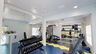 This fitness center offers a comprehensive workout environment designed to accommodate a variety of exercise routines. The space is defined by its bold, eye-catching aesthetic, featuring a striking blue and yellow chevron-patterned floor that energizes the room. An accent wall dressed in horizontal stripes of blue, green, and yellow adds further visual interest and personality to the space.

The layout is thoughtfully organized to maximize functionality. A dedicated free weights area along one wall includes a well-stocked dumbbell rack, with large floor-to-ceiling mirrors extending across that section to enhance the sense of space and allow users to monitor their form. The cardio section is equipped with treadmills and an elliptical trainer, while a multi-function cable and weight machine provides options for strength training exercises.

White walls and crown molding lend a clean, polished finish to the interior, while recessed and surface-mounted lighting ensures the space is well-lit throughout. Ceiling vents support proper air circulation, contributing to a comfortable workout environment. Overall, the fitness center balances practicality with an upbeat, motivating atmosphere.