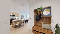 This single-floor residence showcases a thoughtfully designed open-concept layout that seamlessly connects the kitchen, dining, and living areas. The entryway sets an inviting tone with a built-in slatted wood mudroom bench, overhead hooks, and display shelving — a practical yet stylish welcome. Light wood-look flooring flows throughout the main living areas, complementing the warm, neutral palette.

The kitchen is a standout feature, outfitted with flat-front light wood cabinetry, white quartz countertops, a subway tile backsplash, stainless steel appliances, and a generous island with bar seating — ideal for both cooking and casual dining. Recessed lighting and under-cabinet lighting enhance functionality.

The living room is bright and airy, filled with natural light from large windows dressed with sheer curtains and blinds. A glass door provides access to an outdoor balcony area, extending the sense of openness.

The bedroom offers a calm, restful retreat with plush carpeting, ceiling fan, floating wood shelves, and a direct connection to the en-suite bathroom. The bathroom features large-format tile surrounds in the tub/shower combo, wood-toned cabinetry with a white quartz vanity, and wood-look tile flooring, maintaining the cohesive modern aesthetic found throughout the residence.