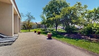 Located at 2150 Wilson Ave in Saint Paul, Minnesota, this outdoor space offers a thoughtfully landscaped grounds area surrounding a multi-story residential building with a stucco exterior. The grounds are defined by a winding paved pathway that curves through the property, bordered by lush native grasses, wildflowers, and dense naturalized plantings that create a vibrant, ecological feel. Mature deciduous trees provide generous shade throughout, contributing to a serene, park-like atmosphere under open Minnesota skies. Large decorative planters with colorful seasonal flowers are positioned along the main walkway, adding visual interest and a welcoming character to the grounds. A designated patio area along the building's facade features lounge chairs positioned beside large windows, offering a relaxing outdoor retreat for residents. Well-maintained lawns stretch along the building's perimeter, accented by flowering perennials including yellow irises and other seasonal blooms. Rock-lined garden beds and tall ornamental grasses add textural variety to the landscape design. The overall ambiance is green, open, and tranquil, with the natural landscaping providing both privacy and a connection to the outdoors throughout the changing seasons.
