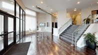 This single-floor space showcases a vibrant, contemporary design anchored by bold yellow accent walls that energize the interior throughout. The main meeting area centers around a sleek glass-top conference table surrounded by modern white chairs, set atop a textured area rug over dark tile flooring. A striking geometric pendant light fixture descends from a curved ceiling detail above, adding architectural interest and focused illumination.

A curated white open-cube shelving unit lines one wall, displaying decorative accessories beneath a series of four coordinating nature-inspired art panels. Adjacent to the conference area, a comfortable lounge nook features gold-and-black striped accent chairs paired with a compact wood side table, offering an inviting informal seating option.

Large floor-to-ceiling glass sliding doors with dark metal frames separate the main area from an adjoining space, allowing natural light to flow freely while maintaining visual connectivity. A dedicated workstation zone accommodates two computer desks and a multifunction printer, making the space fully functional for business operations. The overall ambiance is professional yet welcoming, blending vivid color with polished, modern furnishings.