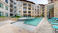 Located at 770 N Eldridge Pkwy in Houston's Energy Corridor, this meticulously designed outdoor amenity space delivers a resort-like experience within a multi-story residential community. The architecture blends stucco and natural limestone masonry, creating a warm, upscale aesthetic that carries throughout the courtyard environment.

At the heart of the space is a generously sized swimming pool with crystal-clear water, complemented by an adjacent raised spa finished in iridescent tile. Broad stone-paved pool decks surround the water features and are furnished with lounge chairs and sun beds, providing ample space for relaxation. Structural limestone columns frame the poolside area, adding architectural character and a sense of enclosure.

The courtyard extends into multiple outdoor living zones, including a shaded dining area outfitted with high-top tables and teal seating beneath vivid blue market umbrellas. A covered outdoor kitchen station houses a stainless steel built-in grill with granite countertops, ideal for social gatherings.

Stone-tiled walkways wind through lush, professionally maintained landscaping featuring palm trees, tropical shrubs, ornamental grasses, and seasonal plantings set within raised stone planters. Gated pedestrian entries with slate tile paths connect the amenity areas to the residential building, ensuring seamless access throughout this thoughtfully planned outdoor retreat.