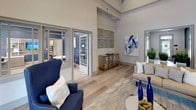Located at 350 Bradley Park Lane in Cumming, Georgia, this single-story community clubhouse offers a refined and welcoming environment designed for both relaxation and productivity. The space is anchored by a dramatic great room with soaring vaulted ceilings and exposed beams, creating an airy, expansive atmosphere throughout. Wide-plank hardwood-style flooring runs seamlessly across the interior, complementing a neutral palette of warm grays and crisp whites.

The main lounge area is thoughtfully furnished with plush seating, layered area rugs, and curated décor including bold blue accent pieces, vibrant wall art, and sculptural metallic wall installations that add a contemporary flair. Tall glass-paneled entry doors flood the space with natural light, while interior transom windows connect the lounge to adjacent rooms without sacrificing openness.

A dedicated business center or conference area is accessible directly from the lounge, featuring workstations and seating suited for meetings or focused work. Additional rooms branch off the central gathering space, offering versatility for various community needs. Lush potted grasses and greenery are strategically placed throughout, bringing a fresh, organic element indoors. Overall, the clubhouse strikes a balance between sophisticated design and functional comfort.