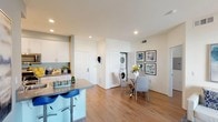 This single-floor residence offers a thoughtfully designed layout that balances comfort and functionality throughout. The open-concept living and dining area is anchored by warm hardwood-style flooring, recessed lighting, and a sliding glass door that invites natural light and provides access to a private outdoor space with views of palm trees and surrounding architecture. The living area offers ample room for lounging and a dedicated workspace, creating versatility for everyday living. Adjacent to the living space, a dedicated dining area features tile flooring and connects seamlessly to the kitchen. The kitchen is well-equipped with stainless steel appliances, including a gas range, built-in microwave, refrigerator, and dishwasher. White cabinetry, gray countertops, and a light blue backsplash accent lend a clean, modern aesthetic, with tile flooring completing the space. A breakfast bar with seating extends the counter space and provides a casual dining option. The bedroom is a comfortable retreat, featuring a sliding glass door with outdoor access and soft neutral tones. A full bathroom includes a wide vanity with white cabinetry, a large mirror, and a shower-tub combination with tile flooring. Completing the unit is an in-unit stacked washer and dryer, conveniently located off the main living area.