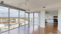 Located in the heart of downtown Durham, NC, this one-bedroom, one-bathroom high-rise residence offers a compelling blend of modern design and sweeping urban panoramas. The open-concept living and kitchen area is anchored by dramatic floor-to-ceiling glass walls that wrap around the corner, flooding the space with natural light and framing breathtaking views of the Durham skyline and beyond. Track lighting overhead complements the abundant daylight, while warm hardwood flooring extends throughout the main living areas, adding a polished, contemporary feel.

The kitchen is well-appointed with sleek white cabinetry, a subway tile backsplash, a full suite of stainless steel appliances including a French door refrigerator, electric range, built-in microwave, and dishwasher, all set against neutral countertops. A sliding glass door leads to a private balcony, where the expansive cityscape stretches to the horizon.

The bedroom continues the high-rise aesthetic with its own floor-to-ceiling windows offering stunning views, plush carpeting, and a ceiling fan. A dedicated closet and hallway provide practical storage. The bathroom features large-format tile flooring, a wide vanity with dark wood cabinetry, white countertops, and a well-lit mirror. The overall ambiance is clean, bright, and distinctly urban.