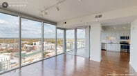 Located in the heart of downtown Durham, NC, this one-bedroom, one-bathroom high-rise residence offers a compelling blend of modern design and sweeping urban panoramas. The open-concept living and kitchen area is anchored by dramatic floor-to-ceiling glass walls that wrap around the corner, flooding the space with natural light and framing breathtaking views of the Durham skyline and beyond. Track lighting overhead complements the abundant daylight, while warm hardwood flooring extends throughout the main living areas, adding a polished, contemporary feel.

The kitchen is well-appointed with sleek white cabinetry, a subway tile backsplash, a full suite of stainless steel appliances including a French door refrigerator, electric range, built-in microwave, and dishwasher, all set against neutral countertops. A sliding glass door leads to a private balcony, where the expansive cityscape stretches to the horizon.

The bedroom continues the high-rise aesthetic with its own floor-to-ceiling windows offering stunning views, plush carpeting, and a ceiling fan. A dedicated closet and hallway provide practical storage. The bathroom features large-format tile flooring, a wide vanity with dark wood cabinetry, white countertops, and a well-lit mirror. The overall ambiance is clean, bright, and distinctly urban.
