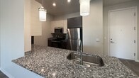 Located at 2176 E Wildermuth Dr in Brighton, Tempe, AZ, this modern single-floor apartment offers 810 square feet of thoughtfully designed living space across 2 bedrooms and 2 bathrooms. The open-concept kitchen and living area serves as the heart of the unit, featuring striking granite countertops with contrasting dark and light cabinetry, stainless steel appliances including a refrigerator, microwave, and range, and stylish pendant lighting over the kitchen island. A tile backsplash and wood-look flooring complete the contemporary aesthetic. The living room is bright and spacious with dark hardwood-style flooring, a ceiling fan, and a sliding glass door opening to a private balcony with outdoor views. Both bedrooms are carpeted and feature ceiling fans and windows with blinds that allow ample natural light. The primary bedroom measures 150 square feet and includes a barn-style sliding closet door. The bathrooms are finished with dark wood vanities and white countertops; one bathroom features a soaking tub with dark tile surround and wood-look flooring. A dedicated closet provides additional storage. The unit's neutral gray palette and modern finishes create a clean, contemporary ambiance throughout.