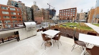 This single-floor amenity space offers a refined, contemporary design tailored for communal gathering and entertainment. The open-plan lounge area serves as the centerpiece, anchored by a dramatic floor-to-ceiling marble fireplace with gold veining, flanked by a wall-mounted flat-screen television. Wide-plank hardwood flooring flows throughout, complementing the neutral, sophisticated palette. Generous floor-to-ceiling windows and glass curtain walls flood the interior with natural light while framing sweeping urban skyline views, creating a seamless connection between indoors and the city beyond.

The fully equipped kitchen features sleek flat-front cabinetry in soft grey and white tones, a bold marble slab backsplash, professional-grade stainless steel appliances including a gas range with a stainless hood, and a large white waterfall island with bar seating. Pendant lighting adds warmth above the island.

Multiple seating arrangements include plush leather sofas, upholstered armchairs in deep burgundy and teal, and intimate conversation groupings suited for both casual relaxation and social events.

The adjoining rooftop terrace extends the entertaining space outdoors, offering a built-in stainless steel grill, patio dining furniture, a lush green lawn area, and unobstructed views of the surrounding urban landscape.