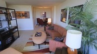 This single-floor space offers an open, flowing layout that connects the living and dining areas in a cohesive, functional arrangement. The living area is anchored by a plush, terra-cotta-toned sofa set accented with decorative throw pillows, complemented by a round upholstered ottoman that doubles as a coffee table. A striped area rug in warm earth tones defines the seating zone, adding texture and visual interest to the carpeted floor. A large potted palm in the corner introduces a natural, organic element, while framed wall art adds personality and color to the neutral, cream-toned walls. Soft, layered lighting from table lamps and a pendant fixture creates a warm and inviting ambiance throughout. The dining area, visible from the living space, features a dark wood table surrounded by cushioned chairs and is illuminated by a pendant light overhead, making it well-suited for intimate meals. The overall design palette leans toward warm browns, rusts, and taupes, creating a unified, comfortable atmosphere. The space feels well-proportioned and thoughtfully arranged, balancing both style and practicality.
