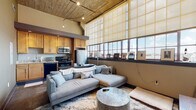 Located at 707 Prudden St in Lansing, Michigan, this single-story loft showcases a compelling blend of industrial heritage and modern design. The open-concept living area immediately draws attention with its dramatically tall, wood-paneled ceiling fitted with track lighting, and a sweeping bank of large factory-style windows that flood the space with abundant natural light and offer open views of the surrounding skyline. The living area comfortably accommodates a generous sectional sofa arrangement centered around a stylish coffee table, creating an inviting gathering space.

The kitchen is efficiently designed with warm maple cabinetry, granite countertops, and a full suite of stainless steel appliances, including a refrigerator, microwave, and electric range, complemented by a dishwasher and undermount sink.

The bedroom is a quiet, enclosed retreat, featuring neutral tones and sufficient space for a full bedroom set. Adjacent to the sleeping area, the bathroom retains the loft's industrial character with an exposed concrete column, polished concrete floors, a walk-in glass-enclosed tile shower with slate-style tile surround, and a modern vessel sink vanity. The entry foyer further highlights the building's raw architectural roots with a prominent exposed concrete column alongside a solid wood door.