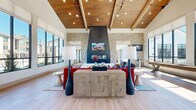 Located in Firestone, Colorado, this single-story community clubhouse on Jake Jabs Boulevard offers a thoughtfully designed gathering space that blends rustic warmth with contemporary sophistication. The main living area commands immediate attention with its dramatic vaulted ceiling clad in warm wood planks, soaring high above light hardwood flooring that flows throughout the space. A floor-to-ceiling stone fireplace serves as the central focal point, flanked by large-format windows that flood the room with natural light and provide views of the surrounding community. Stone columns with dark tile accents frame the transition between zones, lending an architectural richness to the open layout. The lounge area features bold, vibrant furnishings set against the airy backdrop, while oversized windows along one wall create a sun-drenched atmosphere. The dining wing offers a more intimate setting, with a textured geometric ceiling, stylish pendant lighting, and a built-in banquette. Semi-private nook spaces enclosed with glass partitions line the dining area, each accented with vertical wood slat wainscoting and equestrian-themed wall murals, providing cozy spots for smaller gatherings or focused work. Throughout the clubhouse, curated greenery, artistic décor, and a cohesive color palette tie the spaces together into a welcoming, upscale community environment.