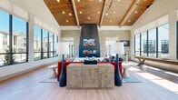 Located in Firestone, Colorado, this single-story community clubhouse on Jake Jabs Boulevard offers a thoughtfully designed gathering space that blends rustic warmth with contemporary sophistication. The main living area commands immediate attention with its dramatic vaulted ceiling clad in warm wood planks, soaring high above light hardwood flooring that flows throughout the space. A floor-to-ceiling stone fireplace serves as the central focal point, flanked by large-format windows that flood the room with natural light and provide views of the surrounding community. Stone columns with dark tile accents frame the transition between zones, lending an architectural richness to the open layout. The lounge area features bold, vibrant furnishings set against the airy backdrop, while oversized windows along one wall create a sun-drenched atmosphere. The dining wing offers a more intimate setting, with a textured geometric ceiling, stylish pendant lighting, and a built-in banquette. Semi-private nook spaces enclosed with glass partitions line the dining area, each accented with vertical wood slat wainscoting and equestrian-themed wall murals, providing cozy spots for smaller gatherings or focused work. Throughout the clubhouse, curated greenery, artistic décor, and a cohesive color palette tie the spaces together into a welcoming, upscale community environment.