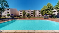 Located in Kenner, Louisiana, this outdoor pool area offers a well-appointed recreational space designed for relaxation and community enjoyment. The centerpiece is a generously sized swimming pool with clear, inviting turquoise water, surrounded by a broad concrete pool deck that provides ample space for lounging. Adjacent to the pool, a decorative brick paver patio adds visual warmth and texture to the outdoor environment, seamlessly connecting the various activity zones.

A dedicated seating area features picnic tables shaded by blue umbrellas, offering a comfortable spot for outdoor dining or socializing beneath mature trees that provide additional natural shade. The entire pool area is enclosed by a black iron fence, ensuring a secure and well-defined perimeter.

A single-story clubroom structure sits alongside the patio, clad in light-colored vinyl siding with a shingled roof. Large sliding glass doors open directly onto the brick paver deck, providing convenient access to the interior and filling the space with natural light. Pool rules are posted on the exterior wall, reinforcing the organized management of the amenity.

The backdrop of a multi-story residential building with balconies frames the space, while lush trees and well-maintained green lawn areas contribute to an open-air atmosphere ideal for outdoor recreation under the sunny Louisiana sky.