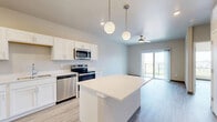 Located at 1439 Boardwalk Ave in Tiffin, Iowa, this one-bedroom, one-bathroom unit offers a thoughtfully designed layout with a clean, modern aesthetic throughout. Light wood-style luxury vinyl flooring runs continuously through the living spaces, complementing the neutral warm-toned walls and crisp white trim.

The kitchen is a standout feature, showcasing white shaker-style cabinetry, light countertops, a full suite of stainless steel appliances — including a refrigerator, range, microwave, and dishwasher — and a central island with pendant lighting overhead. Recessed lighting adds to the bright, functional atmosphere of this space.

The living area benefits from a ceiling fan and sliding glass doors that open onto a private balcony, offering open views of the surrounding Tiffin landscape. A large picture window in the bedroom further enhances natural light and provides elevated views of the neighborhood and beyond.

The bedroom is a well-proportioned retreat with its own closet and continued wood-style flooring. The bathroom is cleanly finished with white cabinetry, a vanity, and matching flooring. A dedicated closet and in-unit utility access round out the practical amenities of this efficiently designed residence.