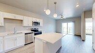 Located at 1439 Boardwalk Ave in Tiffin, Iowa, this one-bedroom, one-bathroom unit offers a thoughtfully designed layout with a clean, modern aesthetic throughout. Light wood-style luxury vinyl flooring runs continuously through the living spaces, complementing the neutral warm-toned walls and crisp white trim.

The kitchen is a standout feature, showcasing white shaker-style cabinetry, light countertops, a full suite of stainless steel appliances — including a refrigerator, range, microwave, and dishwasher — and a central island with pendant lighting overhead. Recessed lighting adds to the bright, functional atmosphere of this space.

The living area benefits from a ceiling fan and sliding glass doors that open onto a private balcony, offering open views of the surrounding Tiffin landscape. A large picture window in the bedroom further enhances natural light and provides elevated views of the neighborhood and beyond.

The bedroom is a well-proportioned retreat with its own closet and continued wood-style flooring. The bathroom is cleanly finished with white cabinetry, a vanity, and matching flooring. A dedicated closet and in-unit utility access round out the practical amenities of this efficiently designed residence.