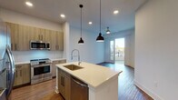 Located at 1050 Slater Rd in Durham, NC, this modern single-floor residence offers a thoughtfully designed open-concept layout with contemporary finishes throughout. The kitchen serves as the heart of the home, anchored by a large center island with a quartz countertop, undermount sink, and sleek pendant lighting. Wood-toned cabinetry pairs with stainless steel appliances — including a French door refrigerator, range, microwave, and dishwasher — to create a functional and stylish cooking space. A subway tile backsplash and recessed lighting complete the look. The living area extends naturally from the kitchen, featuring warm hardwood-style flooring and direct access to a private balcony through glass-paned French doors, bringing in generous natural light. A ceiling fan adds comfort to the space. An in-unit stacked washer and dryer provides added convenience. The bedrooms are finished with plush carpeting and well-proportioned windows that fill the rooms with natural light. One bedroom connects directly to a bathroom featuring warm wood vanity cabinetry, quartz countertops, and a tub-shower combination enclosed by a curved glass door with elegant marble-style tile surround. Throughout the unit, clean white walls, black hardware accents, and wood-look flooring create a cohesive, modern aesthetic.