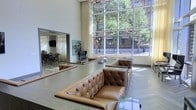 This elegant lobby lounge is defined by its dramatic double-height ceilings and an expansive wall of floor-to-ceiling windows that flood the interior with natural light and frame sweeping views of the surrounding urban streetscape, complete with mature trees and city architecture. The grand scale of the space creates an immediate sense of openness and sophistication upon entry. At the heart of the lounge, a pair of generously sized tufted leather sofas in warm caramel tones are arranged around a low coffee table, offering a refined and inviting seating area. A statement chandelier overhead adds a touch of elegance that complements the clean, contemporary architectural lines. Adjacent to the main lounge, a dining area is visible through an open passage, featuring dark bar-height seating and tables beneath soft, draped curtains and a wall-mounted display screen, suggesting a versatile space suited for social gatherings or events. Artwork adorns the walls, adding color and personality to the otherwise neutral palette of whites and light stone flooring. Together, these elements create a polished, welcoming environment that balances modern design with comfortable, functional living spaces.
