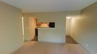 This single-floor space is designed with an open and functional layout that promotes easy flow between living areas. The main living space features wall-to-wall carpeting in a neutral tone, providing a comfortable and cohesive foundation throughout. A central half-wall peninsula separates the kitchen from the living area, creating a pass-through design that maintains visual openness while defining each zone.

The kitchen is equipped with wooden cabinetry, dark countertops, and black appliances, including a refrigerator. The kitchen layout is compact yet practical, with workspace extending along the back wall. A textured ceiling is consistent throughout the space, adding a subtle architectural detail overhead.

Multiple doorways are visible from the main living area, indicating access to additional rooms and hallways. A well-lit corridor leads toward what appears to be a secondary entrance or additional living spaces, enhanced by a ceiling-mounted light fixture. The neutral, beige-toned walls throughout create a clean and understated aesthetic, making the space adaptable to a variety of interior styles. Overall, the layout is straightforward and efficient, ideal for everyday living.