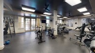 Located at 7105 Old Katy Rd in Houston, TX, this single-floor fitness center offers a comprehensive workout facility designed to support a wide range of exercise routines. The space is thoughtfully organized into distinct zones, each catering to different training needs.

The cardio area is outfitted with multiple treadmills, elliptical trainers, and rowing machines, each positioned near large windows that allow natural light to fill the space and offer views of the surrounding outdoor greenery. Ceiling fans mounted on the bold, dark-painted ceiling contribute to air circulation and add a modern aesthetic contrast to the white structural columns throughout.

A dedicated cycling zone features several stationary spin bikes arranged for group or individual use, with mounted flat-screen televisions nearby for entertainment during workouts. The strength training section is stocked with a full dumbbell rack, medicine balls, a ballet barre, resistance machines, and an adjustable weight bench, catering to both functional fitness and traditional strength training.

Wall-to-wall mirrors in the free weights area enhance the sense of space and assist with form correction. The flooring throughout is durable rubber gym matting, designed for safety and impact absorption. French-style glass doors provide natural light and access points, reinforcing an open, airy atmosphere. This facility is well-suited for a full-body training regimen in a clean, modern environment.