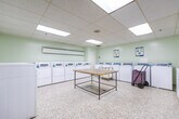 Located at 177 Nonquon Rd in Oshawa, Ontario, this shared laundry facility spans a single floor and is organized into three distinct functional areas. The space features light green walls, durable speckled terrazzo flooring throughout, and a suspended acoustic tile ceiling equipped with recessed fluorescent lighting that ensures consistent, well-lit conditions across all sections.

The main laundry area is generously sized and lined with rows of coin-operated front-load washers and dryers along the perimeter walls, providing ample capacity for multiple users simultaneously. A central folding table offers a convenient surface for sorting and folding laundry, while rolling laundry carts are available for transporting loads with ease. Baseboard heating units run along the walls to maintain comfort year-round.

A dedicated waiting area adjoins the laundry zone, furnished with metal bench seating and a wall-mounted fan for ventilation. Informational notices are posted on the walls, and a payment terminal is conveniently accessible near the entrance corridor. A recycling bin is also present, reflecting a commitment to responsible waste management.

The transitional corridor connecting the sections provides clear sightlines throughout the facility, enhancing ease of navigation. Overall, the space is designed with practicality and functionality in mind, offering a clean, organized, and efficiently laid-out communal laundry environment.