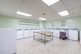 Located at 177 Nonquon Rd in Oshawa, Ontario, this shared laundry facility spans a single floor and is organized into three distinct functional areas. The space features light green walls, durable speckled terrazzo flooring throughout, and a suspended acoustic tile ceiling equipped with recessed fluorescent lighting that ensures consistent, well-lit conditions across all sections.

The main laundry area is generously sized and lined with rows of coin-operated front-load washers and dryers along the perimeter walls, providing ample capacity for multiple users simultaneously. A central folding table offers a convenient surface for sorting and folding laundry, while rolling laundry carts are available for transporting loads with ease. Baseboard heating units run along the walls to maintain comfort year-round.

A dedicated waiting area adjoins the laundry zone, furnished with metal bench seating and a wall-mounted fan for ventilation. Informational notices are posted on the walls, and a payment terminal is conveniently accessible near the entrance corridor. A recycling bin is also present, reflecting a commitment to responsible waste management.

The transitional corridor connecting the sections provides clear sightlines throughout the facility, enhancing ease of navigation. Overall, the space is designed with practicality and functionality in mind, offering a clean, organized, and efficiently laid-out communal laundry environment.