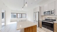 Located at 345 St Peter St in downtown Saint Paul, MN, this modern one-bedroom, one-bathroom unit offers a thoughtfully designed layout with contemporary finishes throughout. The open-concept kitchen is a standout feature, showcasing two-tone cabinetry — warm wood lower cabinets paired with crisp white uppers — complemented by a quartz countertop island with striking wood base, stainless steel appliances, tile backsplash, and stylish pendant lighting. Light-toned hardwood flooring flows through the main living and kitchen areas, adding warmth and continuity. The spacious bedroom features plush carpeting and a wide bank of dark-framed windows that invite generous natural light and urban views of the surrounding cityscape. Two closets serve the bedroom area, including a well-appointed walk-in closet outfitted with multi-tiered wire shelving for versatile storage. The bathroom is sleek and modern, featuring a wood-grain vanity with a white countertop, matte black fixtures, and a walk-in shower. Additional conveniences include an in-unit stacked washer and dryer. The overall aesthetic is clean and contemporary, with bright white walls and natural wood accents creating a cohesive, inviting atmosphere throughout.