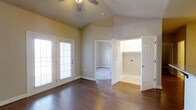 This single-floor residence offers a thoughtfully designed layout centered around a bright and open main living area. The space immediately draws attention with its vaulted ceilings, which lend a sense of spaciousness and airiness throughout. A ceiling fan is mounted at the peak, providing both function and a subtle architectural accent.

Rich, dark hardwood flooring runs throughout the main living area, adding warmth and a polished aesthetic to the space. Neutral, warm-toned walls complement the flooring and create a welcoming ambiance that adapts well to a variety of interior styles.

Natural light flows generously into the living area through a large multi-pane glass door fitted with horizontal blinds, offering both brightness and privacy control. This door also provides direct access to an outdoor area, seamlessly connecting interior and exterior spaces.

From the main living area, multiple doorways lead to additional rooms. One opens to a carpeted bedroom or secondary room, while another — fitted with a classic six-panel white door — reveals a hallway or utility area with additional access points. White trim and baseboards throughout contribute to a clean, cohesive finish. Overall, the space combines practical layout with comfortable, inviting design details suited for everyday living.