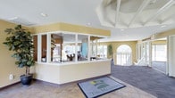 The Meadows is a two-story facility that makes a strong first impression with its inviting, light-filled entrance. The lobby welcomes visitors with a prominent reception desk enclosed in glass panels, offering staff clear sightlines across the space while maintaining an open, accessible feel. The flooring transitions from warm tile at the entry to plush carpet in the surrounding areas, subtly defining different zones within the space. Tall, vaulted ceilings with a skylight overhead amplify the sense of openness and draw in generous amounts of natural light. Warm yellow accent walls complement the white trim and neutral tones throughout, creating a cheerful and professional atmosphere. Arched windows and wide-pane glass features along the perimeter frame views of the outdoors, reinforcing the connection to the surrounding environment. Beyond the reception area, the interior opens into broader communal spaces furnished for gathering and conversation, with additional greenery adding a natural touch to the décor. A staircase with white railings provides access to the second floor, which expands the facility's overall footprint. The overall design balances functionality with a welcoming ambiance, making The Meadows a comfortable and well-organized environment for visitors and staff alike.