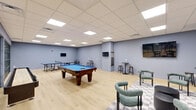 This single-floor recreational room offers a generously sized, open-plan layout designed specifically for leisure and social engagement. The space is finished with light hardwood flooring and cool-toned, blue-gray walls that lend a clean, modern aesthetic throughout.

At the heart of the room sits a full-size pool table with a vibrant blue felt surface, accompanied by a dedicated cue rack mounted on the wall nearby. A ping-pong table positioned toward one end of the room provides additional gaming options. Multiple flat-screen TVs are wall-mounted at various points, ensuring clear sightlines from across the space.

A casual lounge area is defined by a checkered area rug, anchoring a comfortable sofa, rounded accent chairs in muted teal upholstery, and a pair of cylindrical coffee tables. High-top tables paired with bar stools are distributed around the perimeter, offering informal seating for spectators or casual conversation.

The drop ceiling is outfitted with a grid of recessed panel lighting that evenly illuminates the entire room. A striking steel-framed glass partition wall separates this game room from an adjacent space, adding an industrial-modern design element while maintaining visual openness. The overall ambiance is energetic yet comfortable, making this room well-suited for both competitive play and relaxed social gatherings.
