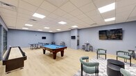 This single-floor recreational room offers a generously sized, open-plan layout designed specifically for leisure and social engagement. The space is finished with light hardwood flooring and cool-toned, blue-gray walls that lend a clean, modern aesthetic throughout.

At the heart of the room sits a full-size pool table with a vibrant blue felt surface, accompanied by a dedicated cue rack mounted on the wall nearby. A ping-pong table positioned toward one end of the room provides additional gaming options. Multiple flat-screen TVs are wall-mounted at various points, ensuring clear sightlines from across the space.

A casual lounge area is defined by a checkered area rug, anchoring a comfortable sofa, rounded accent chairs in muted teal upholstery, and a pair of cylindrical coffee tables. High-top tables paired with bar stools are distributed around the perimeter, offering informal seating for spectators or casual conversation.

The drop ceiling is outfitted with a grid of recessed panel lighting that evenly illuminates the entire room. A striking steel-framed glass partition wall separates this game room from an adjacent space, adding an industrial-modern design element while maintaining visual openness. The overall ambiance is energetic yet comfortable, making this room well-suited for both competitive play and relaxed social gatherings.