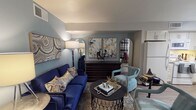 Located at 1600 S Valley View Blvd in Las Vegas, Nevada, this single-floor space presents a thoughtfully curated interior with a distinctly contemporary design sensibility. The living area immediately captures attention with its rich color palette, anchored by a bold navy blue sofa adorned with decorative pillows featuring metallic and earth-toned accents. A plush teal accent chair adds a complementary pop of color, creating an elegant and cohesive visual dynamic throughout the space.

The room is appointed with carefully selected furnishings, including a sleek round black coffee table styled with decorative objects, books, and a metallic star accent piece. Arched gold-framed mirrors flank a central diptych botanical wall art arrangement, adding depth and artistic character to the cool blue-gray walls. A large abstract canvas in muted tones anchors one wall, further enriching the layered aesthetic.

Warm ambient lighting from tall floor lamps enhances the inviting atmosphere, while a patterned area rug grounds the seating arrangement. Glimpses of adjacent areas reveal plaid drapery and additional furnishings, suggesting a well-appointed and cohesive floor plan throughout. The overall ambiance is sophisticated, warm, and carefully composed, reflecting a designer sensibility suited to comfortable yet stylish everyday living.