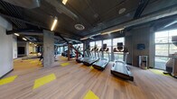 Located at 25 Charlton Street in Everett, Massachusetts, this fitness center delivers a well-equipped, modern workout environment designed with an industrial aesthetic. The space is defined by exposed concrete columns, open ductwork overhead, and dark ceiling treatments that lend it a raw, urban character. Linear LED lighting runs throughout, ensuring the facility is well-lit at all times.

Large multi-pane windows line the perimeter walls, flooding the floor with generous natural light and offering views of the surrounding outdoors — a standout feature that enhances the overall workout experience. The flooring is a combination of light wood-tone material accented by bold yellow rectangular inlays, adding energy and visual structure to the open layout.

The cardio section is prominently featured, housing a full row of treadmills, elliptical machines, StairMasters, and rowing machines, all positioned to take advantage of the natural light from the expansive windows. Beyond cardio, the space extends into a strength and functional training zone equipped with cable machines, free weights, and additional resistance equipment visible along mirrored walls that amplify the sense of space. The generous, open floor plan allows for comfortable movement throughout, supporting a full range of fitness routines in a dynamic and motivating environment.