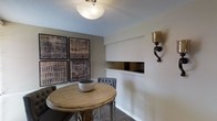 This single-floor space showcases a carefully curated interior with a strong emphasis on texture, tone, and artistic detail. The dining area serves as a central focal point, featuring a round natural wood table paired with upholstered gray chairs that add a refined, contemporary touch. The warm wood tones of the table contrast elegantly with the neutral taupe wall coloring that runs throughout the space.

A standout feature of the dining area is the gallery wall arrangement of four large-format framed artworks, each displaying intricate blue-and-beige abstract patterns that lend a sophisticated, artistic character to the room. Complementing these pieces is a recessed niche built directly into the wall, finished in a reflective gold-toned interior, which serves as both a decorative and functional display element.

Wall-mounted sconce lighting with an ornate wrought iron bracket and a hammered gold shade adds a classic, artisanal quality to the space. Overhead, a frosted glass flush-mount ceiling fixture provides soft, diffused illumination. The textured ceiling and hardwood flooring further contribute to the layered, tactile quality of the interior. Overall, the space presents a cohesive blend of rustic and contemporary design sensibilities.