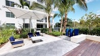 This elegant outdoor courtyard offers a well-appointed retreat designed for both relaxation and social gathering. The space is anchored by a large, light-toned paver patio that provides a clean, polished foundation throughout the area. Towering palm trees rise prominently from carefully maintained landscaping beds filled with lush tropical greenery, creating a vibrant, resort-like atmosphere. A large cantilever umbrella offers shaded seating over a grouping of modern wood-frame chairs upholstered in rich navy blue cushions, arranged around a sleek, low-profile white table — ideal for casual lounging or conversation. Bold, geometric royal blue planters serve as striking architectural accents, adding a pop of color that complements the navy furnishings. The surrounding landscaping blends ornamental shrubs, smooth decorative gravel, and tropical foliage to frame the space with a polished, manicured aesthetic. A warm wood deck border transitions seamlessly from the paved patio, adding natural texture and warmth to the overall design. The backdrop features a modern white multi-story building with open balconies and large glass openings, while wrought iron fencing and lush perimeter greenery complete the sense of an enclosed, private oasis. Abundant natural light and clear blue skies enhance the open, airy ambiance of this thoughtfully designed outdoor space.
