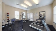 This fitness center is designed to accommodate a well-rounded workout routine within a comfortable, single-floor layout. The space features neutral beige walls, dark rubber flooring ideal for exercise, and flush fluorescent lighting that ensures consistent illumination throughout. Multiple windows fitted with white blinds allow natural light to filter in, contributing to an open and airy atmosphere.

The cardio section is equipped with a treadmill, elliptical trainer, and recumbent stationary bike, providing a variety of low- and high-impact aerobic options. A multi-station cable weight machine supports a range of strength training exercises, while a medicine ball rack stocked with multiple weighted balls adds functional fitness versatility. Additional strength equipment and a flat workout mat round out the available training tools.

A framed fitness center rules posting is mounted near the entrance, and a small utility station along one wall provides added convenience. The space is accessed through a set of French doors with louvered shutters, which also allow views to an adjoining area. An emergency exit door provides secondary access. The overall design is functional and clean, creating a practical environment suited for daily fitness use.