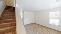 This two-floor space is designed with functionality and accessibility in mind. The layout, as revealed by the 3D model, features a central staircase with a handrail connecting both levels, providing straightforward access between floors. The stairwell opens to a well-lit landing with a panel door entry point.

Throughout the space, rooms are finished with neutral beige carpeting and crisp white walls, creating a clean, understated aesthetic. Multiple rooms on both floors feature several windows dressed with white blinds, allowing for generous natural light and outdoor views of surrounding greenery. One bright room is highlighted by a pendant light fixture, adding a focal point to the space.

The kitchen is equipped with warm oak cabinetry spanning the full perimeter, offering ample storage. White appliances, including a range, refrigerator, and dishwasher, are included, and the room features tile flooring and a stainless steel sink beneath a window.

Two bathrooms serve the space. One features a tub-shower combination with chrome fixtures, while the other is designed for accessibility, equipped with grab bars positioned near the toilet and along the walls, alongside a wide-open vanity with a large mirror and a multi-bulb light fixture above.