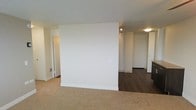 This single-floor space is characterized by its clean, minimalist design and practical layout. A prominent partial wall serves as a natural divider, separating two distinct zones within the unit. The carpeted area, finished in a neutral beige tone, offers a softer, more comfortable feel, while the adjoining section transitions to dark hardwood flooring, adding contrast and a polished aesthetic.

The walls throughout are finished in crisp white, contributing to a bright and open atmosphere. Ceiling-mounted track lighting in the hardwood-floored section provides focused illumination, complementing the flush ceiling light fixture nearby. White baseboards run along the perimeter, adding a clean, finished look to the space.

Doorways visible in the background suggest access to additional rooms or storage areas, hinting at a functional floor plan. The partial wall features a standard electrical outlet, indicating its use as a practical interior partition rather than a purely decorative element. Overall, the space presents a neutral, move-in-ready environment with a straightforward design well-suited for a variety of furnishing arrangements.