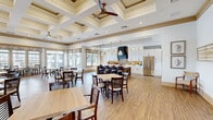 Located at 1111 W Bay Ave in Barnegat, NJ, this expansive single-floor community clubhouse spans a generously sized footprint designed to accommodate a variety of gathering and sales functions. The grand entrance foyer greets visitors with soaring double-height ceilings, textured grasscloth-style wall coverings, warm wood flooring, and elegant wall sconces, establishing a refined coastal aesthetic throughout. A dedicated sales office is prominently situated near the entry, complemented by a literature display and comfortable tufted seating in teal.

The heart of the space is a showcase kitchen featuring floor-to-ceiling white shaker cabinetry, a bold navy range hood, a gold-toned herringbone tile backsplash, stainless steel appliances, and a rustic wood-and-navy island with pendant lighting overhead. Adjacent to the kitchen, a spacious dining area accommodates numerous tables and chairs beneath an impressive coffered ceiling with ceiling fans, while large windows along the perimeter flood the room with natural light and offer outdoor views.

An elevator provides convenient access between levels, and the space also includes a wide covered outdoor terrace finished with herringbone brick pavers, a warm wood ceiling, and white columned railings overlooking the surrounding community — ideal for outdoor gatherings and socializing.