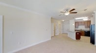 Located at 485 Traebert Circle in Raleigh, NC, this single-floor residence offers an open and airy layout designed for comfortable everyday living. The main living area is finished with wall-to-wall carpet in a neutral tone, contributing to a warm and inviting atmosphere throughout the space. Crown molding runs along the ceiling perimeter, adding a refined architectural detail that elevates the overall aesthetic.

A ceiling fan with an integrated light fixture is centrally positioned in the living area, providing both air circulation and ambient lighting. The walls are painted in a soft, neutral beige tone that enhances the sense of openness and pairs well with the light-colored trim and white panel doors visible throughout.

The living space flows naturally toward the kitchen area, which is partially visible and features pendant lighting hanging above what appears to be a countertop or breakfast bar with a stone or granite surface. The kitchen's lighter cabinetry and pendant fixtures provide a subtle contrast to the adjoining living space.

Multiple interior doors are visible toward the rear of the unit, suggesting dedicated spaces for bedrooms or additional rooms, all consistent with the clean, neutral design palette carried throughout the home.