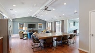 The Event Room at The Abbys At Inverness in Birmingham, Alabama, is a well-appointed, single-floor gathering space designed to accommodate a variety of social functions. The room's most notable architectural feature is its vaulted, beam-accented ceiling, which rises prominently over the central lounge area and is fitted with dual ceiling fans and recessed lighting, contributing to an open, airy atmosphere throughout.

Rich hardwood flooring runs continuously across the space, lending warmth and cohesion to the distinct functional zones within the room. A central conference-style dining area features a substantial solid wood table surrounded by modern upholstered chairs, making it well-suited for group meetings or communal dining. Adjacent to this, a comfortable lounge area is furnished with leather sofas, a coffee table, and a wall-mounted television set atop a media console, with framed artwork adding visual interest to the slate-blue accent wall.

Large sliding glass doors and floor-to-ceiling windows line the perimeter, flooding the interior with natural light and providing direct views of the surrounding landscaped grounds. A kitchenette area offers granite countertops, warm wood cabinetry, open shelving, and a microwave, providing practical support for catered events. The overall ambiance is modern, welcoming, and thoughtfully designed for community use.